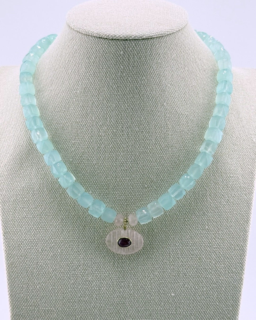 Blue chalcedony faceted cube strand necklace with a carved rose quartz oval pendant set with a ruby, on a linen bust