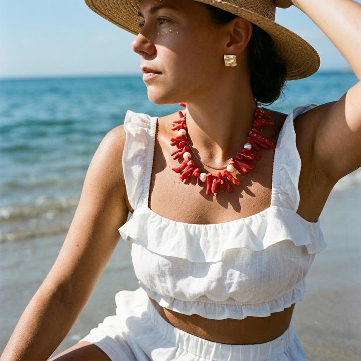 woman in a white ruffled top and straw hat on the beach, wearing the red coral and pearl statement necklace